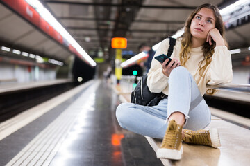 Young attractive girl in jeans waiting for subway train on a platform, using her smartphone