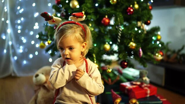 Little girl with a festive headband babbles while sitting near a decorated Christmas tree