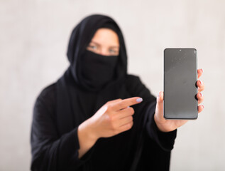 young Muslim girl with her face covered by burka shows empty dark smartphone screen. Close-up, gray...