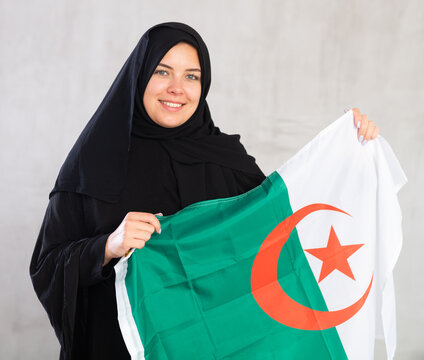 smiling Muslim woman in traditional black hijab holds flag of Algeria. Portrait of female Muslim with Algerian flag on gray background