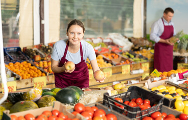 Polite friendly saleswoman standing at counters in greengrocery store, holding ripe red apples...