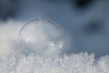 A small transparent soap bubble lies on fresh snow crystals. The background is blue. There is space...
