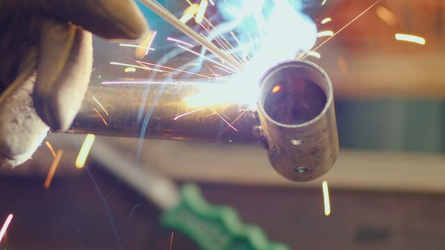 Hand in protective glove welding metal pipe details together with welding torch producing flame, fume and sparks. Close-up view, focus of foreground
