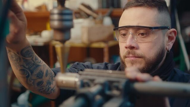 Young bearded man with tattoos wearing safety glasses drilling metal workpiece with spiral step bit in the repair shop. Rack focus, close-up view