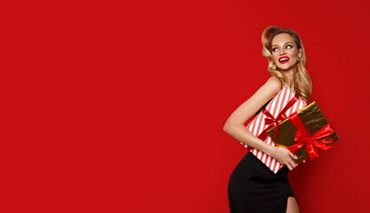 Beautiful young white blonde woman in black dress holds gold gift box with bow on red background....