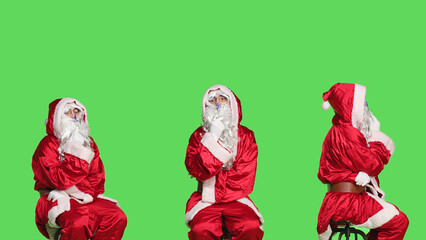Santa on chair brainstorm gift ideas sitting over greenscreen backdrop, thinking about presents before christmas eve holiday celebration. Young man dressed as saint nick in studio.