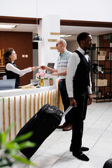 Female concierge assists elderly man at front desk, providing smooth check-in process and verifying reservations. African american bellboy helps retired senior male guest with suitcase to hotel room.