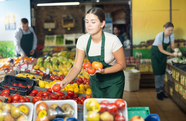 Young woman seller of fruit and vegetable store works near showcase with garden-stuff, sorting tomato, checking goods.