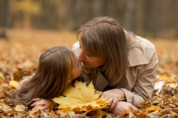 Young beautiful mother and her little cute daughter lying on the fall leaves and have fun in the autumn park. Concept of parenting and carefree childhood.