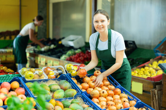 Positive skilled saleswoman in green apron working in fruit and vegetable section of supermarket, laying out ripe pomegranates on display stand..