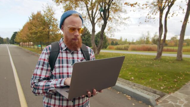 Stylish male in yellow sun glasses, checkered shirt with backpack walking along deserted alley in beautiful autumn park, surfing Internet on silver laptop. High quality 4k footage