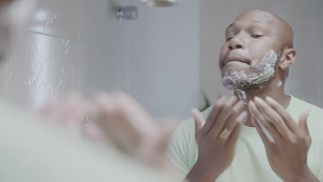 Black man applying shaving foam on his face in front of the mirror. Cinematic 4k.
