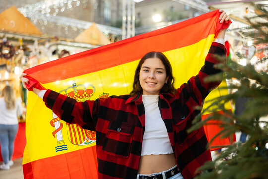 Smiling young traveler girl in white crop top and warm red and black plaid shirt enjoying walk on traditional street Christmas fair, waving national flag of Spain..