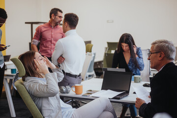 Obraz premium Young, blonde, female employee suffering from migraine during working hours with her cross generational team at modern office space. She massages her head due to the intense pain.