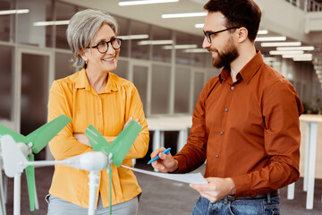 Obraz premium Team of smiling business partners engineers, developers talking, planning startup project, cooperation using windmill working in modern office. Alternative, wind energy, innovation concept