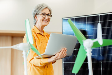 Smiling senior woman, engineer, developer using laptop standing near solar panel and windmill...