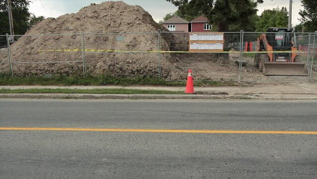 big pile of dirt behind silver fence with building permits on bulletin board on it with yellow black construction vehicle parked next to it, road in front vehicles passing and residential homes behind