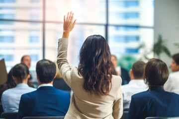 Rear view of a casual businesswoman raising her arm in a full conference room waiting for her turn to speak up - obrazy, fototapety, plakaty
