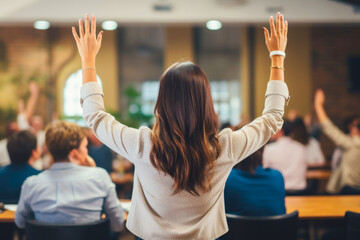 Rear view of a casual businesswoman raising her arms in a full conference room waiting for her turn to speak up - obrazy, fototapety, plakaty