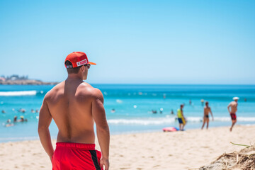 Rear view of a male lifeguard who dutifully watches over swimmers ready to take immediate action in case of a life threatening situation in the ocean - obrazy, fototapety, plakaty