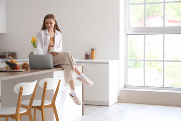 Happy young woman holding cup of coffee and sitting on table with modern laptop in light kitchen