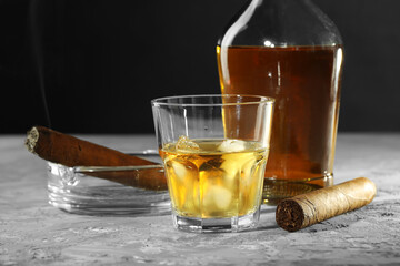 Bottle, glass of whiskey with ice cubes and cigars on grey table