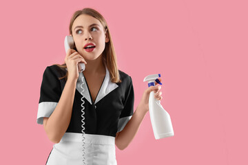 Beautiful female janitor with bottle of cleaning product talking by mobile phone on pink background