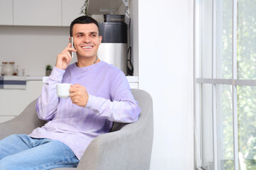 Handsome young man drinking coffee while talking by mobile phone and sitting on armchair near window