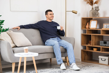 Handsome young man with cup of coffee sitting on sofa in modern living room