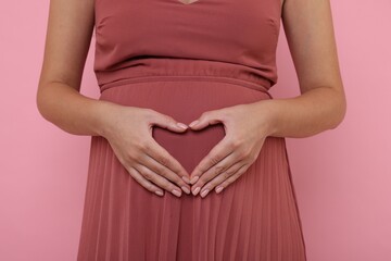 Pregnant woman in dress making heart with hands on pink background, closeup