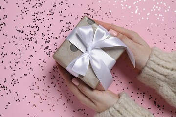 Christmas present. Woman holding gift box and confetti on pink background, top view