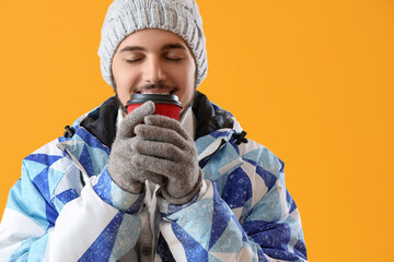 Young man in winter clothes drinking hot coffee on yellow background, closeup