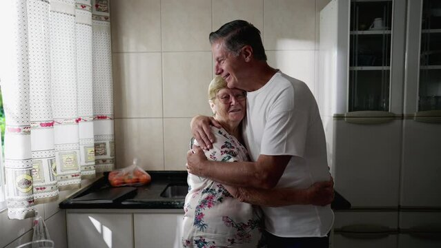 Adult son embracing elderly mother standing at home kitchen. Authentic real people lifestyle. Man interacting with senior woman, family values care-taking for senior people