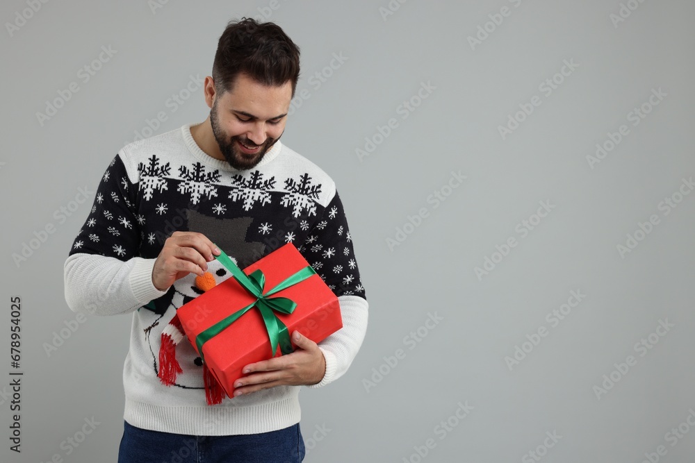 Wall mural young man in christmas sweater opening gift on grey background. space for text