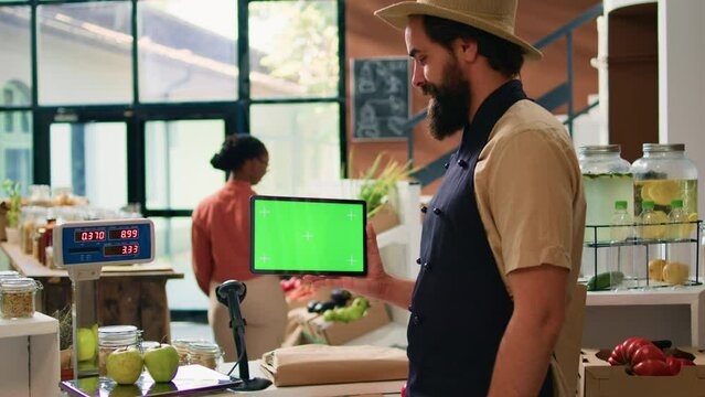 Storekeeper holds tablet with greenscreen in zero waste eco supermarket, using copyspace mockup on mobile gadget standing at checkout counter. Small business owner presents isolated display.