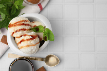 Scoops of tasty ice cream with mint, candies and caramel sauce on white tiled table, flat lay....