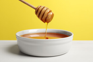 Pouring tasty honey from dipper into bowl at white wooden table against yellow background, closeup