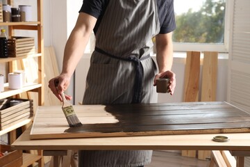 Man with brush and can applying wood stain onto wooden surface indoors, closeup