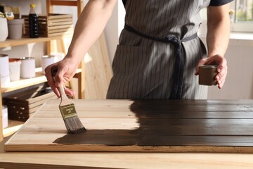 Man with brush and can applying wood stain onto wooden surface indoors, closeup