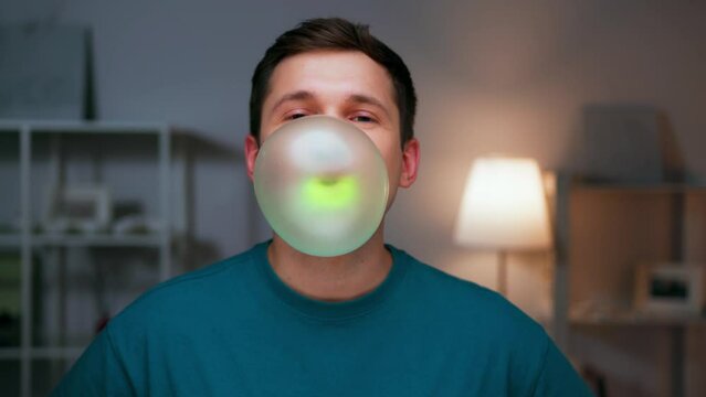 Portrait of Young Man Chewing Bubble Gum and Looking at Camera Close Up. Joy of Modern Person with Bubblegum in Mouth Making Colorful Ball. Alone Guy in Room Loving and Enjoying Bright Green Balloon