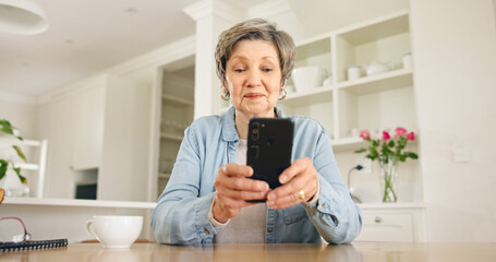 Senior woman typing on smartphone in home at table for social media, reading email or chat. Elderly...