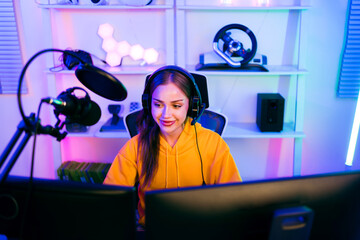 Young Caucasian woman professional gamer in yellow hoodie and headphones sit on a chair and gaming table, pc, keyboard, monitor, microphone. Prepare for competition, cast gameplay or record a podcast.