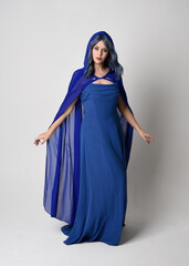 Naklejka premium Full length portrait of beautiful female model wearing elegant fantasy blue ball gown and flowing cape with hood. Standing pose, with gestural arms reaching out . Isolated on white studio background.