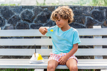 Cute diverse little boy eating a snow cone or shave ice outdoors on a public street bench. Enjoying a cool refreshment on a hot summer day