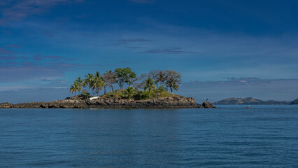 Naklejka premium A beautiful tropical island in the blue ocean. Palm trees grow on the rocks. Tiny silhouettes of people and boats are visible. A mountain range in the distance against the azure sky and clouds.