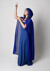  Full length portrait of beautiful female model wearing elegant fantasy blue ball gown, flowing cape with hood. Standing pose walking away, gestural arms reaching out. Isolated on studio background