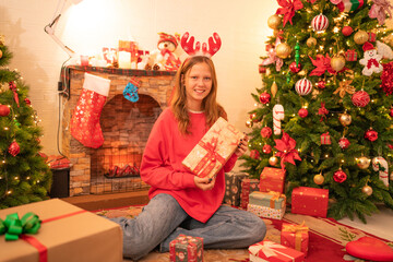 Obraz na płótnie Canvas Happy girl holding christmas gift box with smile hild wearing Chrismas jumper posing on floor near fireplace and xmas tree.