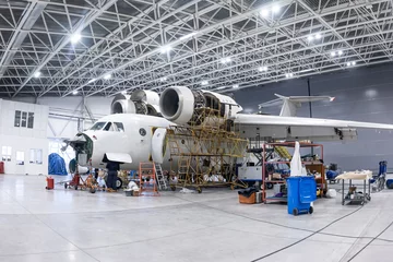 Zelfklevend Fotobehang White transport aircraft in the aviation hangar. Airplane under maintenance. Checking mechanical systems for flight operations © Dushlik