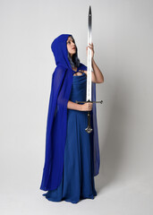 Full length portrait of beautiful female model wearing elegant fantasy blue ball gown, flowing cape with hood.
Standing pose walking away, holding a sword weapon Isolated on white studio background.