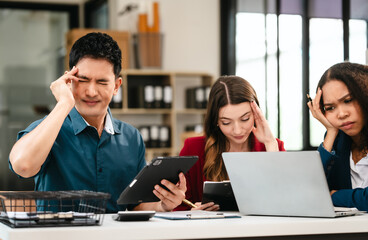 Business team meeting with stressed man rubbing his forehead, while two women review documents, appearing serious and focused.